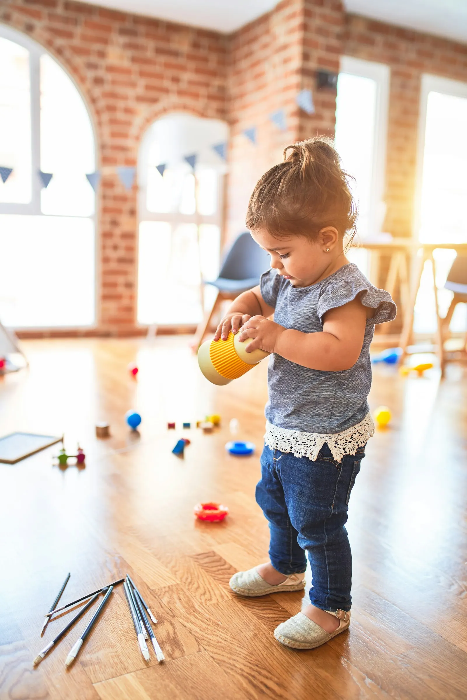 Une petite fille tient une tasse jaune dans une salle de jeux ensoleillée, entourée de jouets
